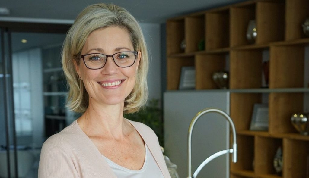 Blonde woman wearing glasses smiles in front of fitted units at a Schmidt Kitchens showroom.