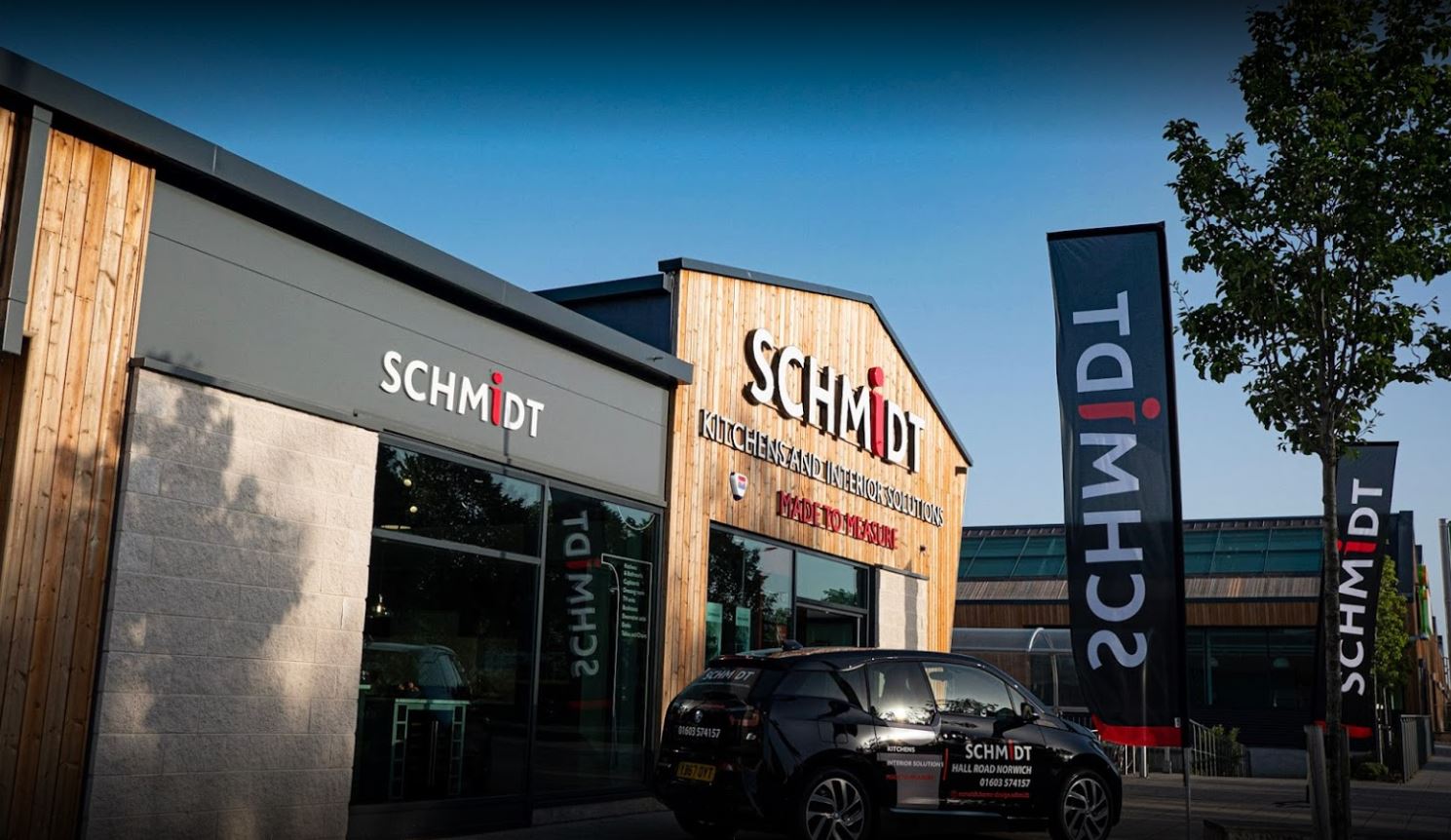 An exterior shot of a Schmidt Kitchens building with wooden cladding, branded car, and marketing flags. This image captures showroom branding and outdoor promotional assets.