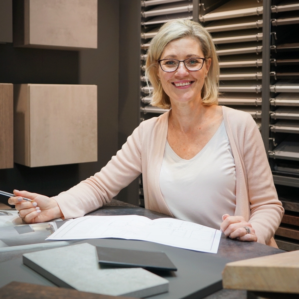 A smiling kitchen designer sits at a showroom table surrounded by colour and surface samples. She helps clients explore materials and layout options for their new kitchen.
