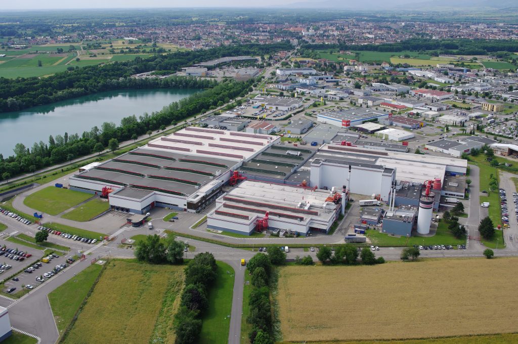 Large-scale manufacturing plant with parking, surrounded by countryside and town. Demonstrates the company’s industrial scale and European production capabilities.