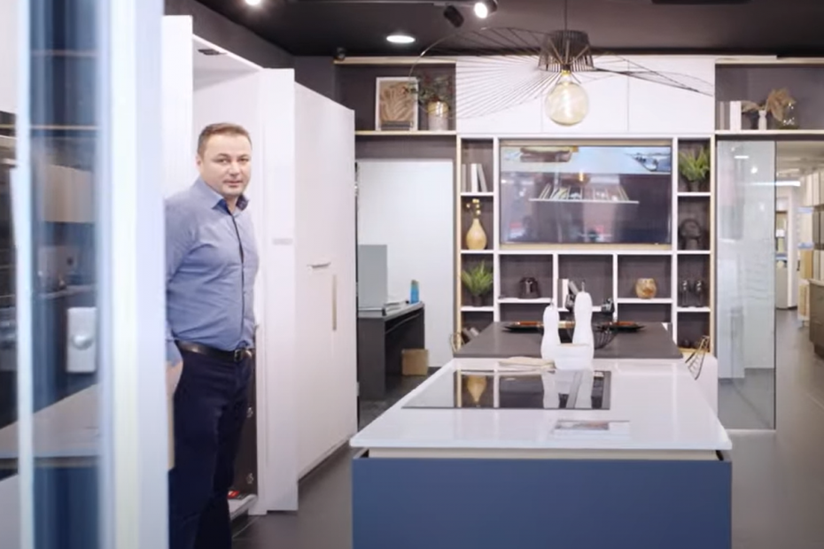 A showroom designer stands beside a sleek kitchen island with white worktops and black induction hob. The room is styled with decorative shelving and accent lighting.