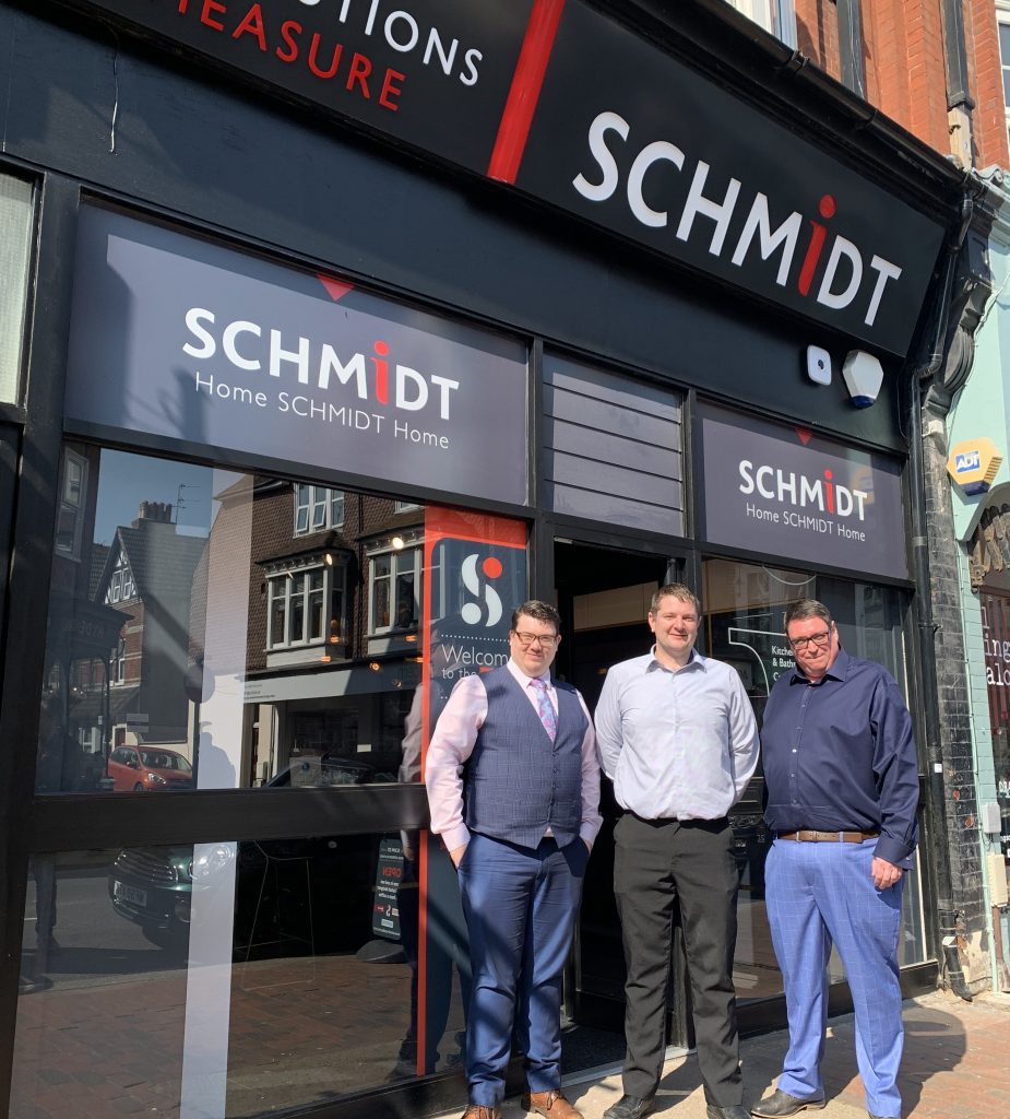 Three team members stand proudly outside a Schmidt showroom, dressed smartly to welcome visitors. The bold signage and urban backdrop highlight the brand's high-street presence.