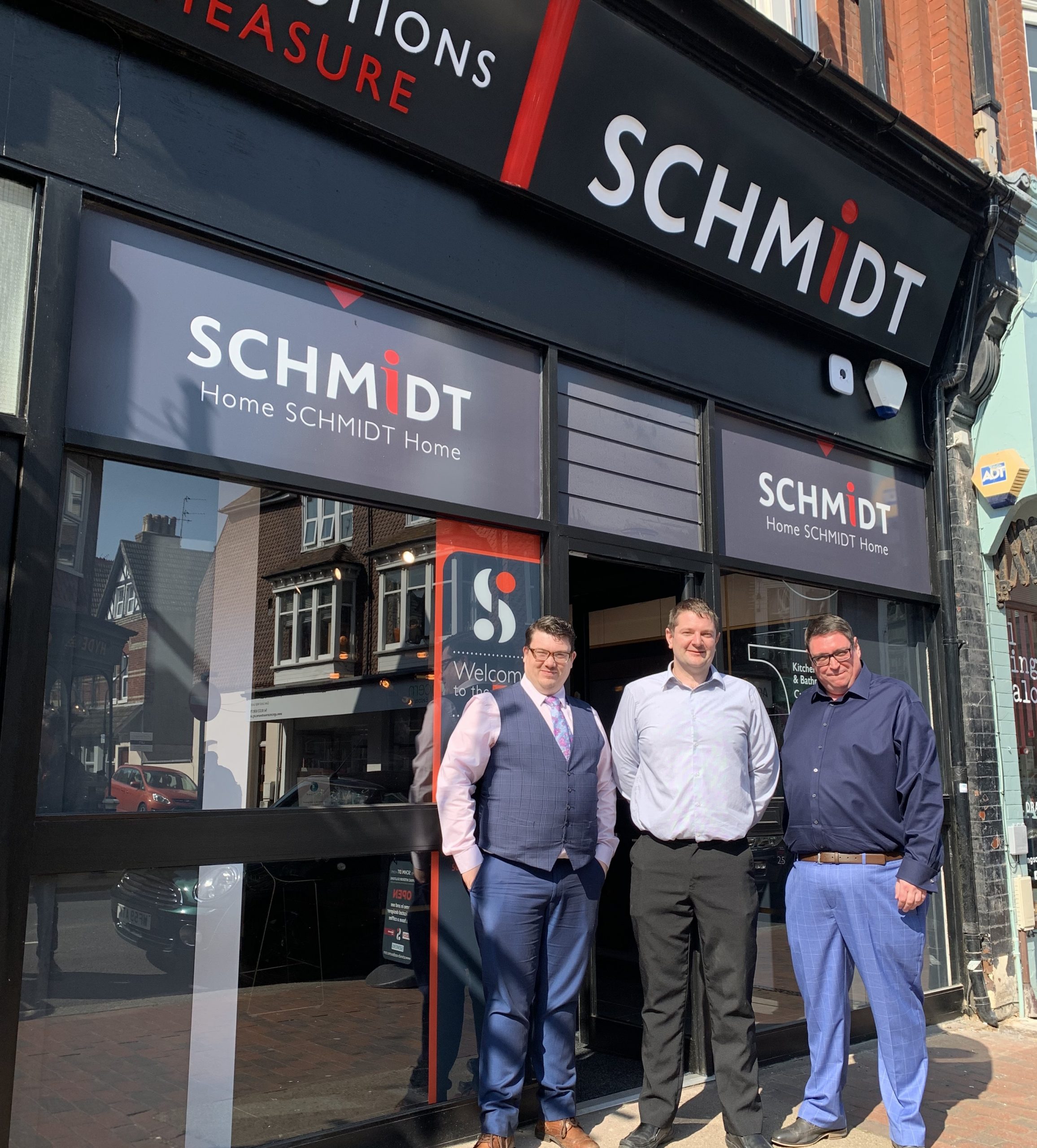 Three team members stand proudly outside a Schmidt showroom, dressed smartly to welcome visitors. The bold signage and urban backdrop highlight the brand's high-street presence.