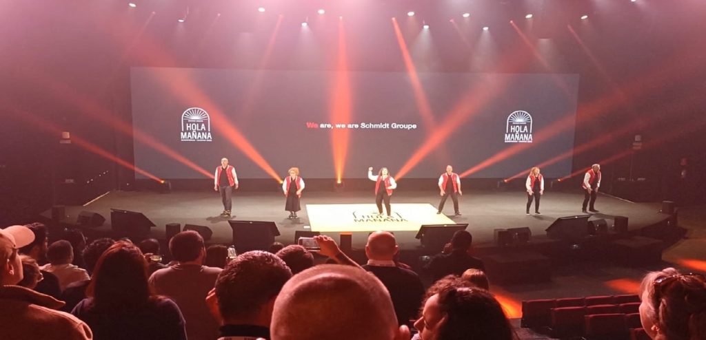 Schmidt Groupe team performing on stage at Hola Mañana event. A group of employees in red jackets stands under dramatic lighting in front of a branded screen and cheering audience.