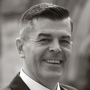 Scott Hardy Black and white portrait of a smiling man in a suit. Professional headshot featuring a clean background and confident expression.