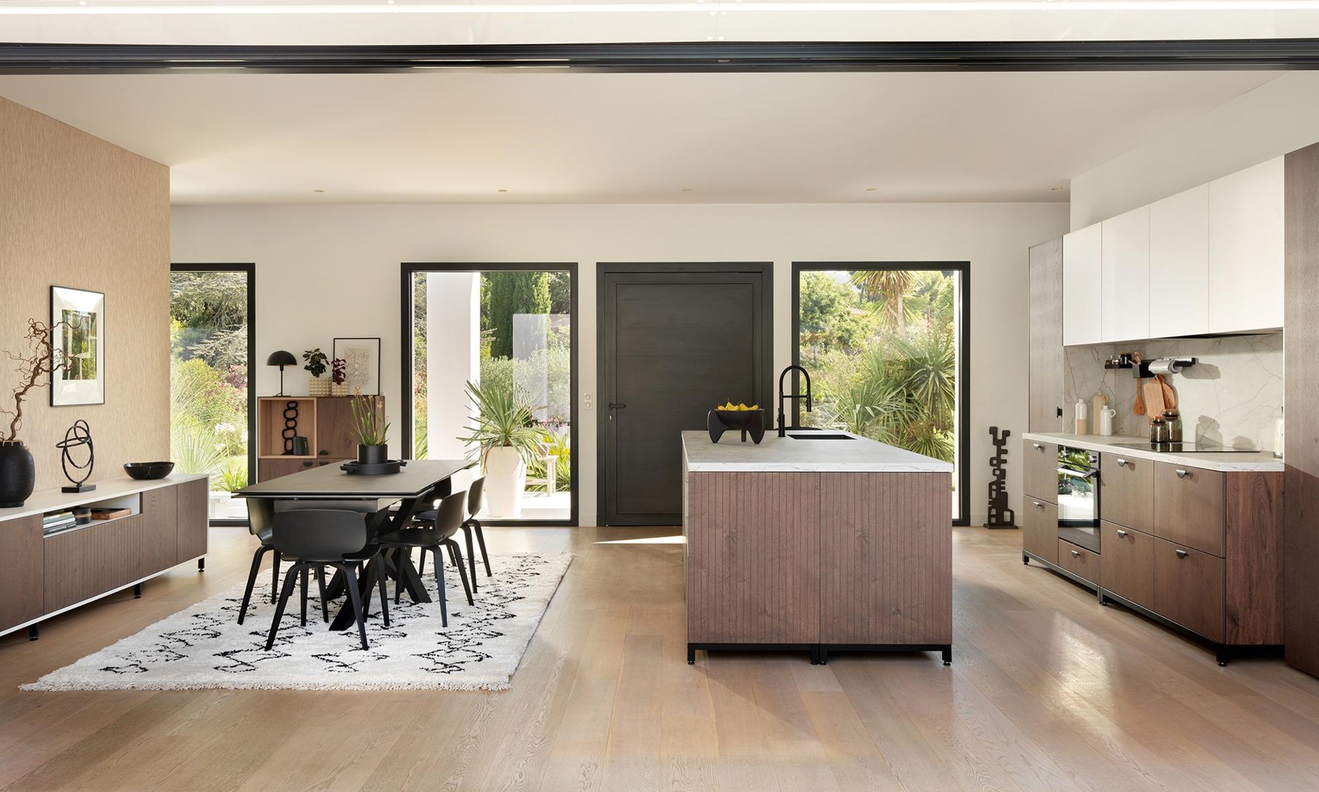 Open-plan kitchen-diner with wood finishes and black framed windows. A bright, modern space featuring natural textures, central island, and direct garden access.