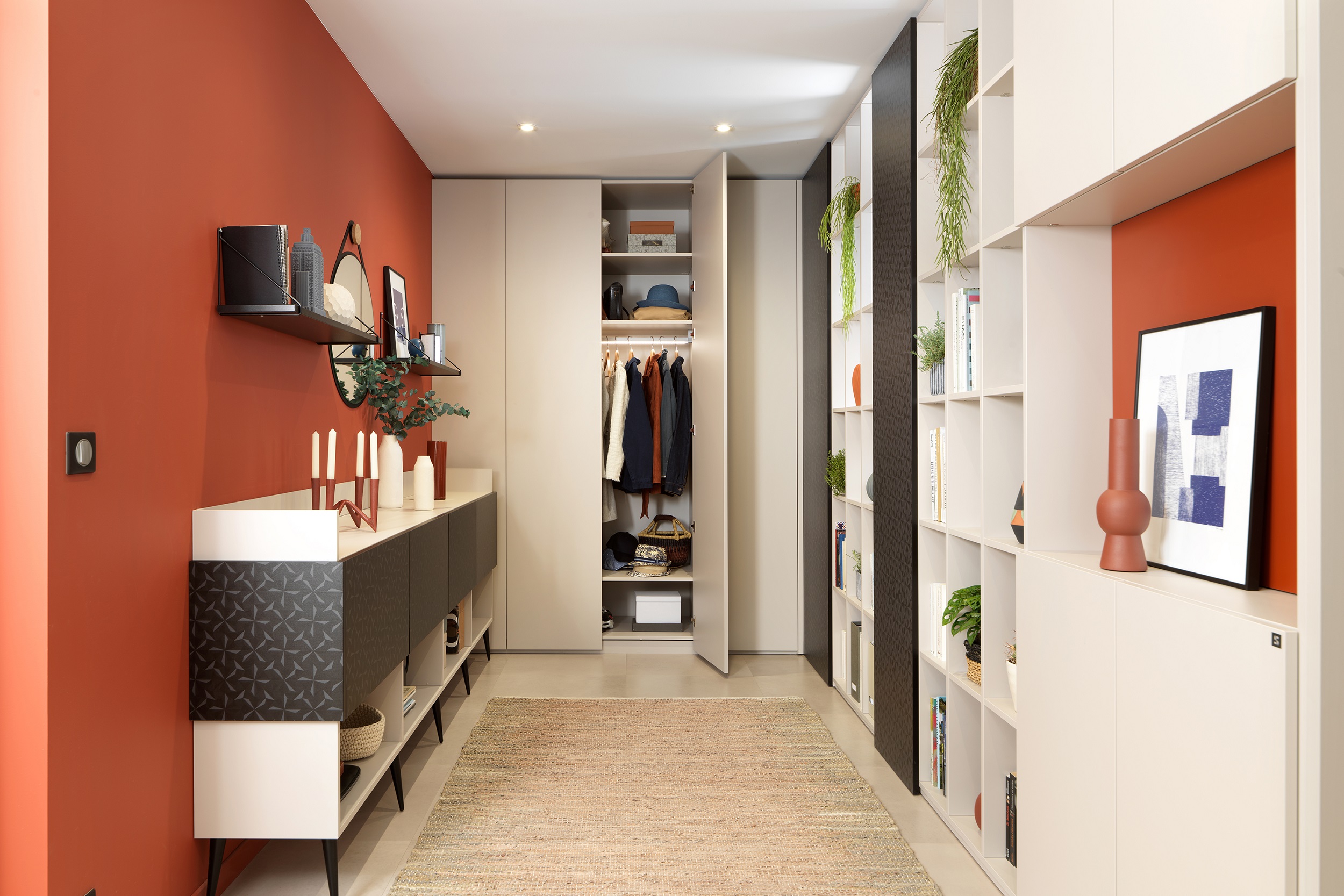 A modern hallway with a bold red feature wall and sleek built-in shelving units filled with books and decorative items. The space includes a patterned sideboard, circular mirror, and a neutral runner rug.