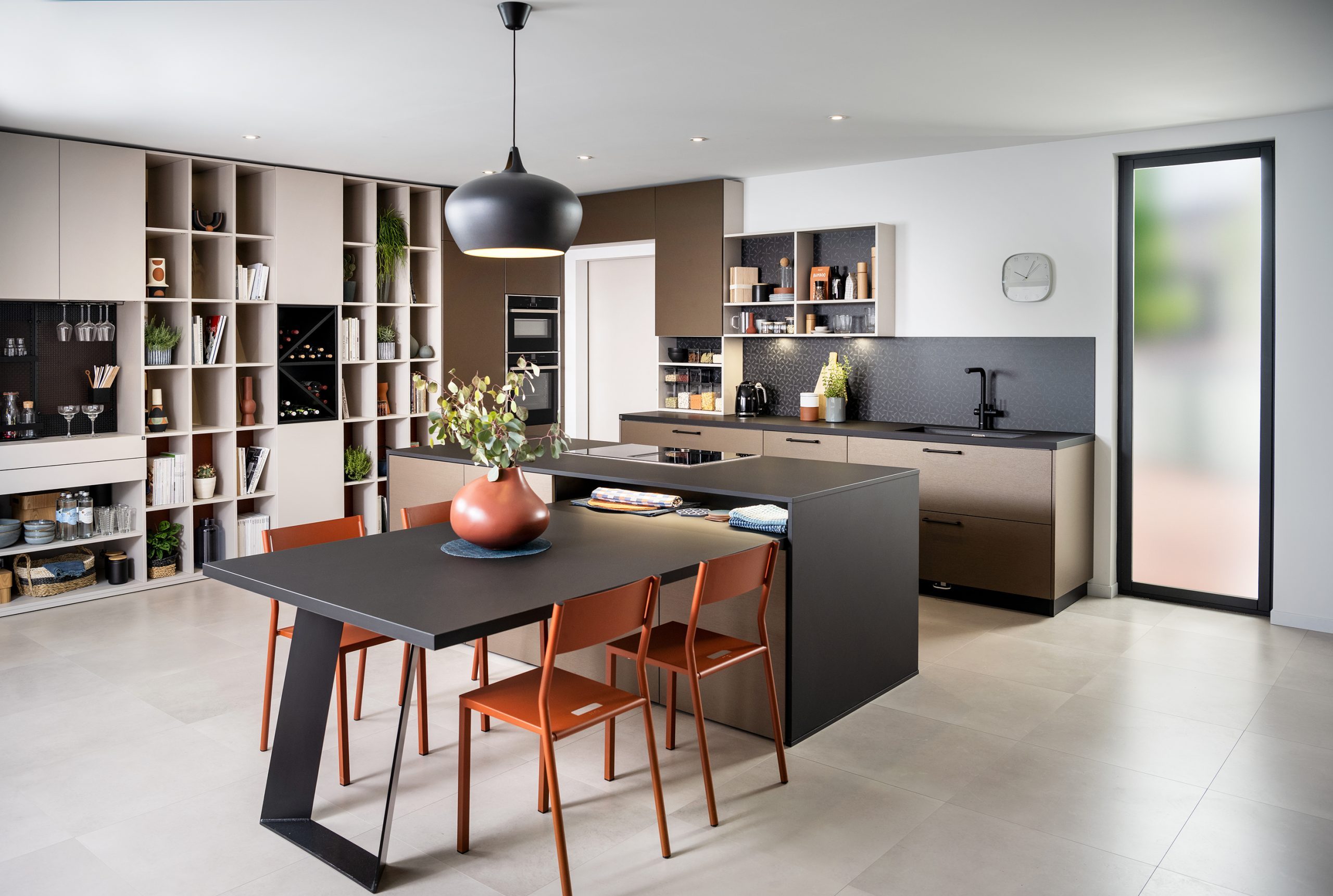 A contemporary kitchen with integrated open shelving, dark cabinetry, and burnt orange chairs surrounds a central island. Decorative plants and tableware add character and warmth to the clean layout.