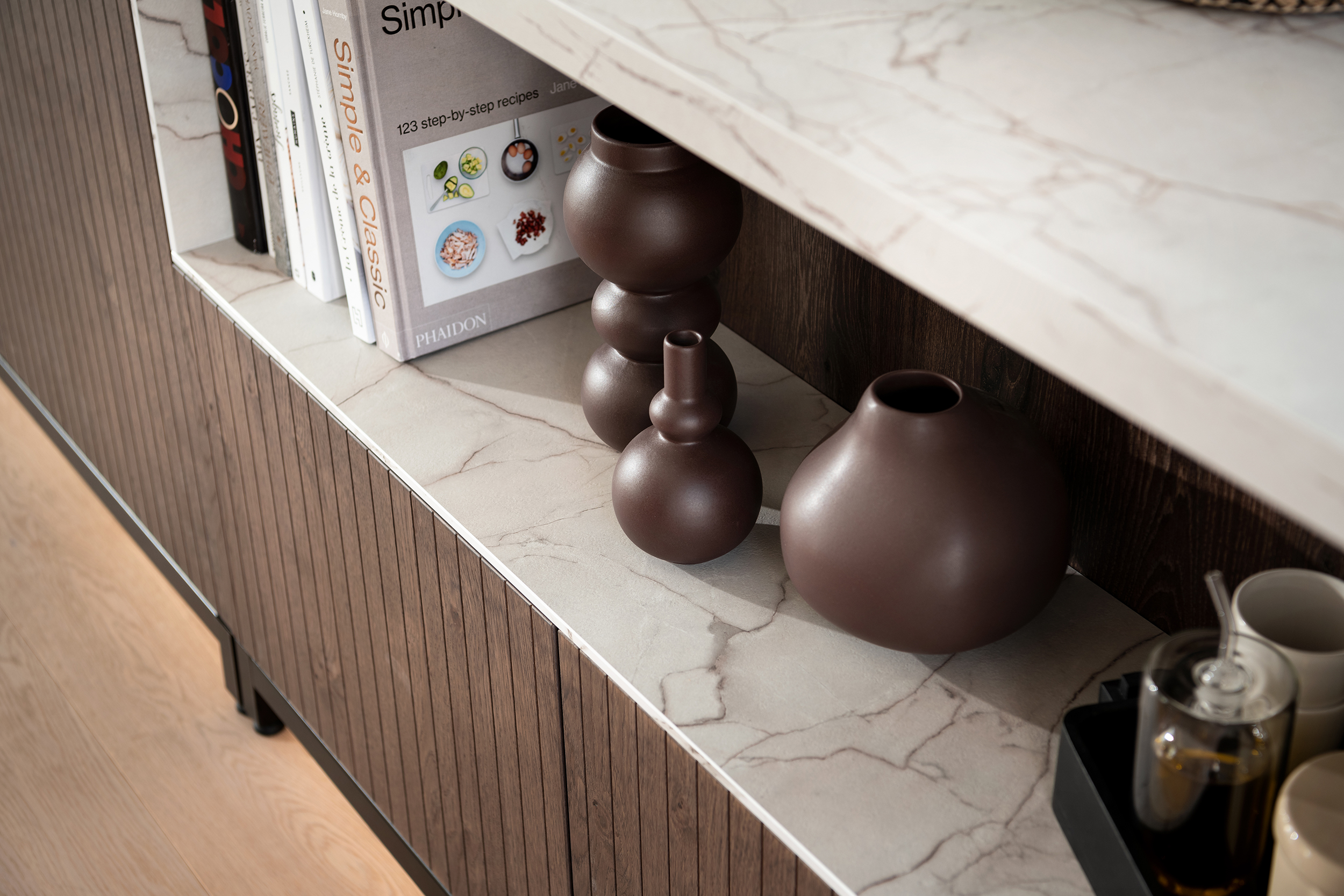 A close-up of a kitchen shelf reveals matte ceramic vases in various organic shapes. The surrounding shelf has a faux-marble backing that ties into the kitchen island surface.
