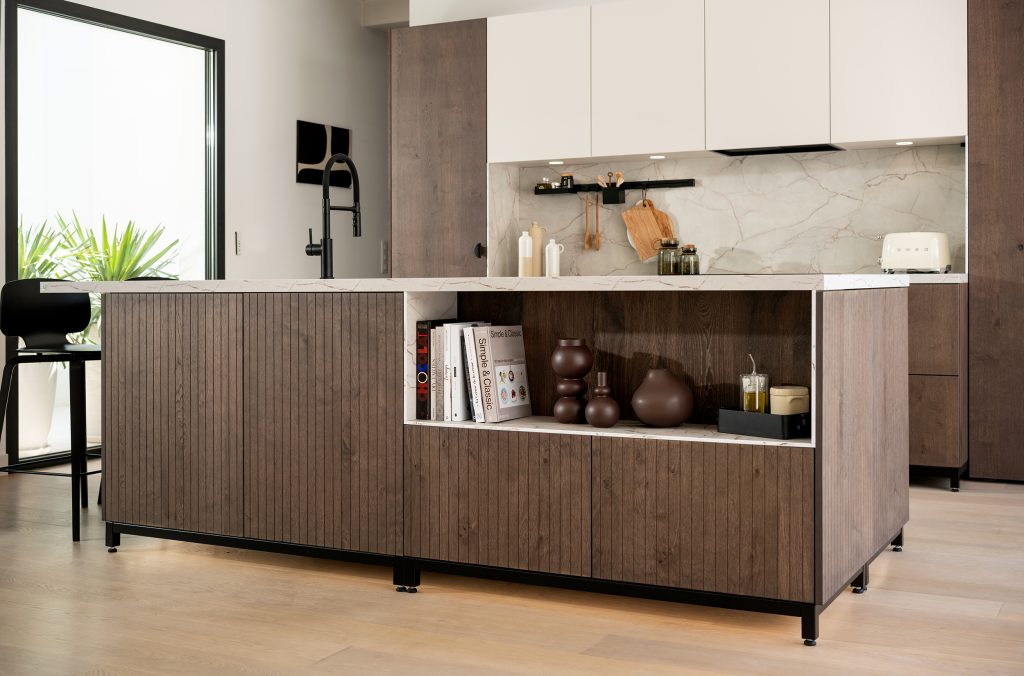 The close-up shows a timber-panelled island with a marble-effect surface and open shelving filled with books and decorative vases. The texture and colour contrast highlight the mix of materials used in the design.