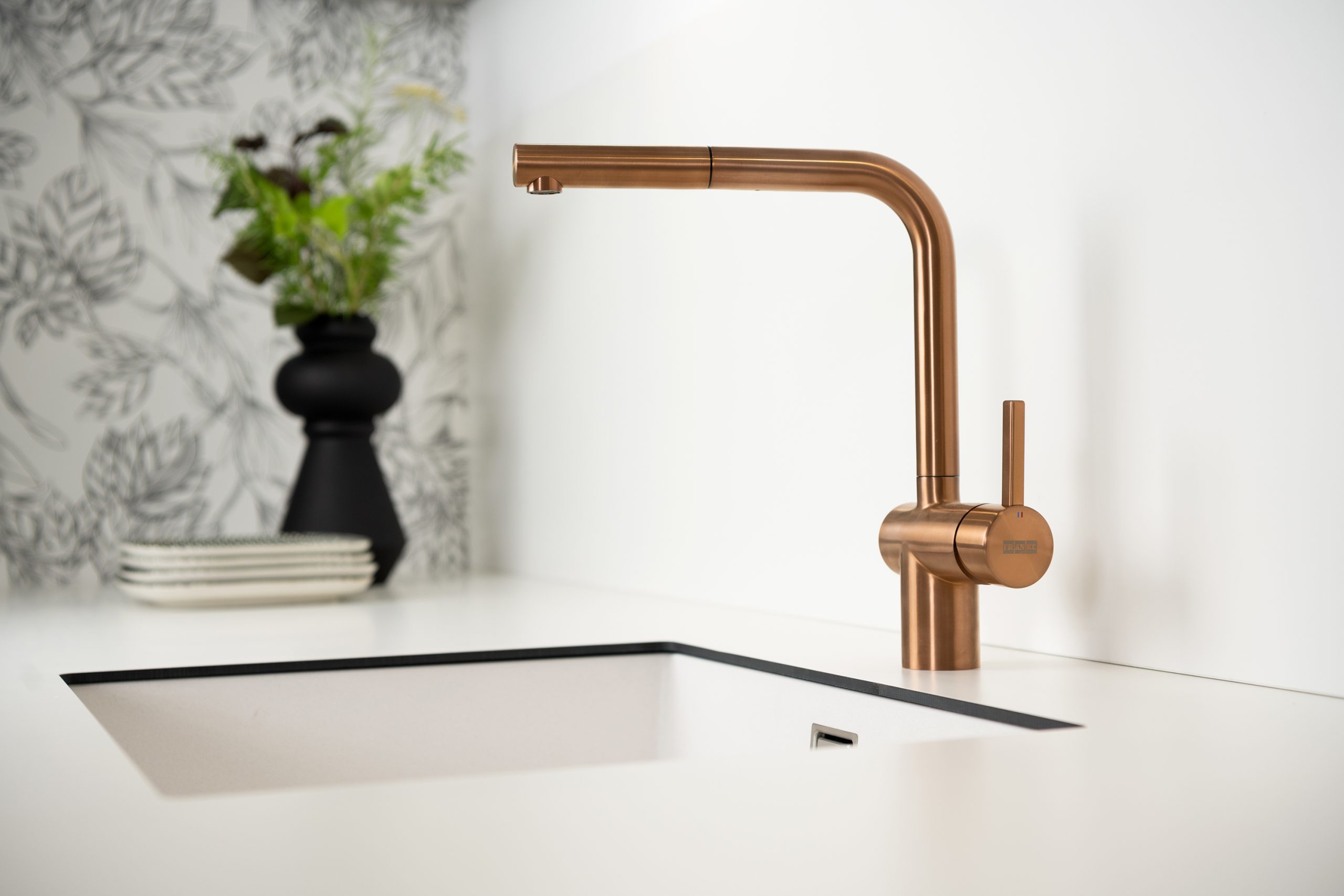 A minimalist kitchen tap in brushed copper stands out against a white countertop. A floral black vase and stacked plates sit in the blurred background near a patterned feature wall.