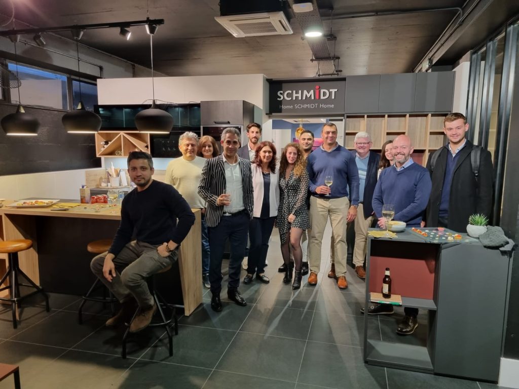 A group of people gather inside a Schmidt showroom, standing together in a relaxed setting with drinks and kitchen displays in the background. The lighting is warm and the space is styled with modern cabinetry.