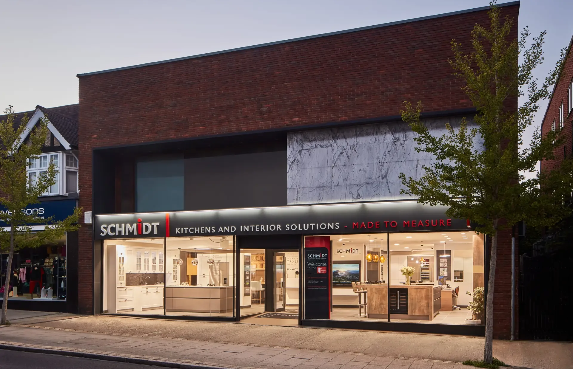 A Schmidt Kitchens showroom with illuminated signage and full-height glass windows showcasing elegant interior solutions. The sleek storefront design reflects premium kitchen and home interiors.
