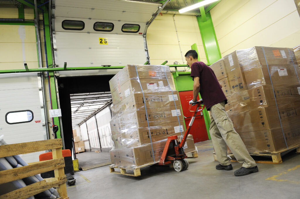 Warehouse loading area with packed pallets. A worker moves shrink-wrapped boxes onto a loading dock with a red pallet jack inside a distribution centre.
