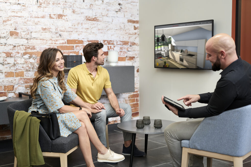 A couple views a digital kitchen layout with help from a consultant in a showroom setting. Wall-mounted display and product samples enhance the experience.