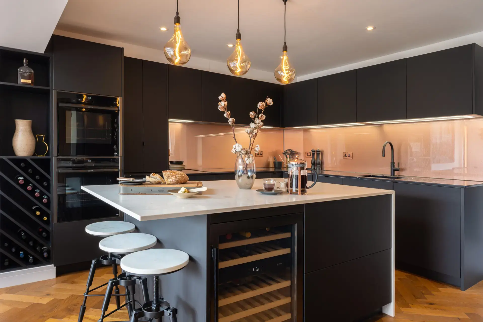 A sleek black kitchen with built-in oven, wine rack, and pendant lighting over a central island. Copper accents and soft lighting enhance the modern, upscale atmosphere.