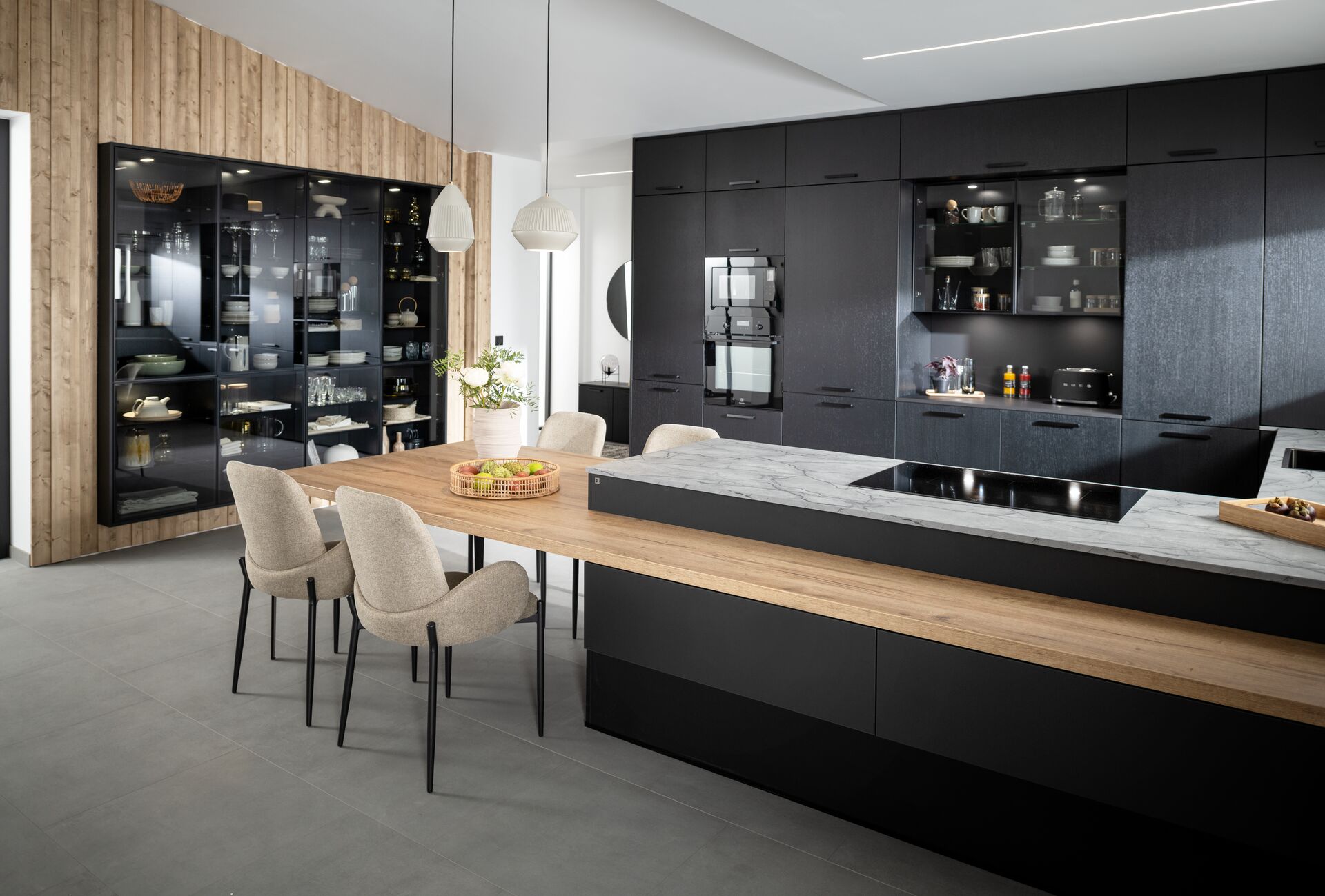 This modern kitchen combines black cabinetry with a dual-surface island made from marble and wood. Backlit glass cabinets showcase premium dishware, adding elegance and depth.